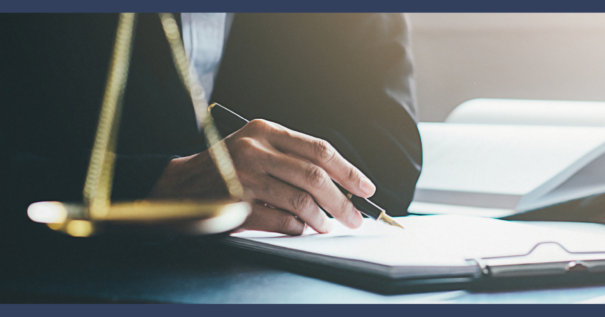 Man in a suit signing legal paperwork, justice scales on desk to his side