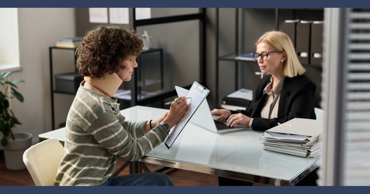 Lady at desk wearing a neck split, sitting opposite a lay who is wearing glasses and a suit while typing notes into a computer